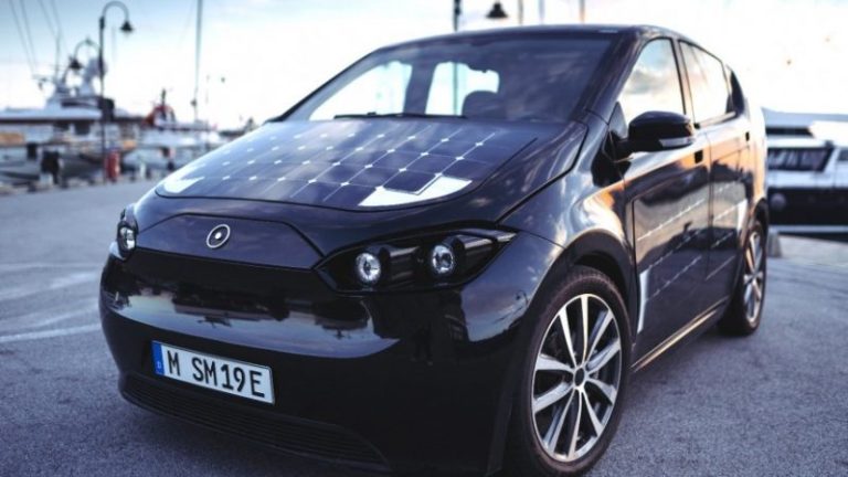 A black electric car with solar panels on its hood is parked near a waterfront marina, with boats and streetlights visible in the background.
