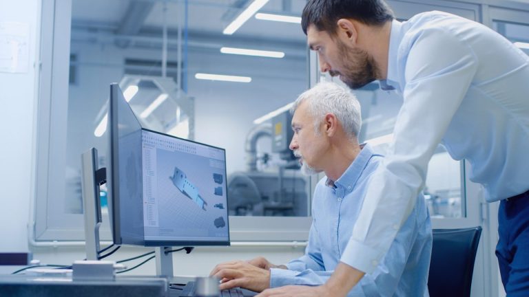 Two men in a modern office work together at a computer displaying a 3D CAD model of a mechanical part; one man is seated using the computer, while the other stands and looks at the screen.