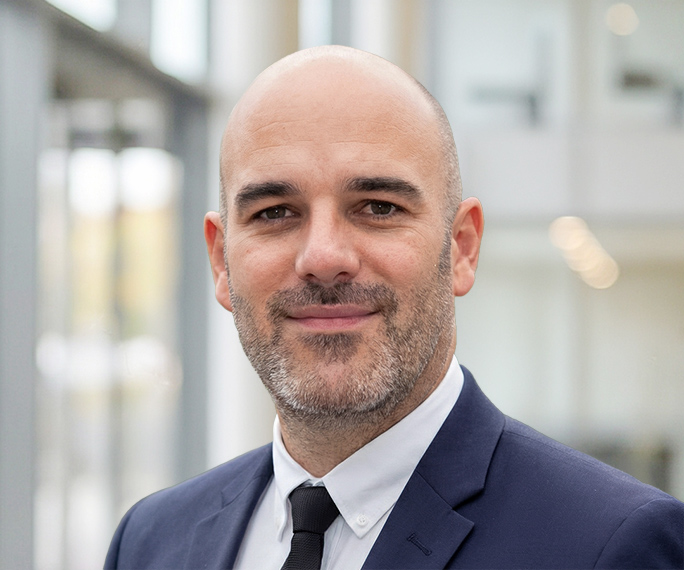 A smiling bald man with a short beard, wearing a dark suit, white shirt, and black tie, stands in a modern, bright office setting with large windows and blurred background.