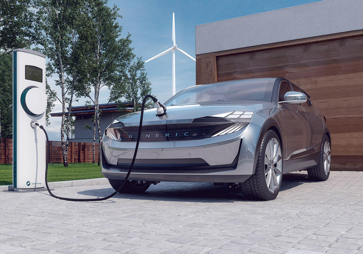 A silver electric car is charging at an outdoor charging station on a paved driveway. A modern house and trees are in the background, and a wind turbine is visible in the distance.