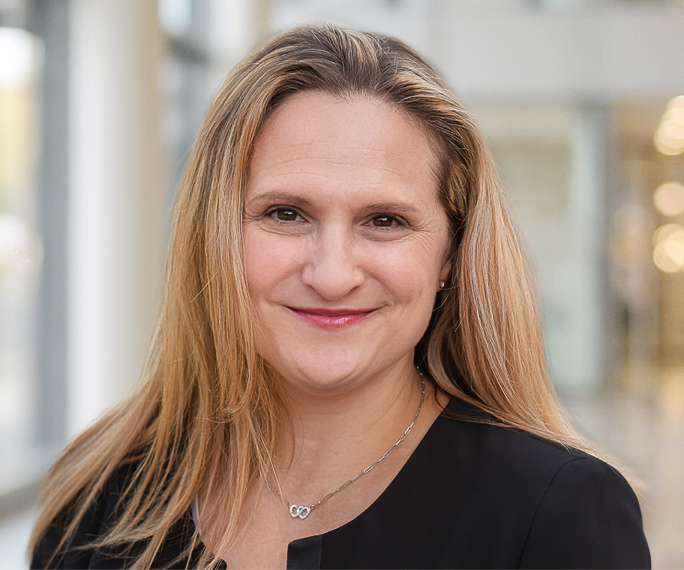 A woman with long blonde hair wearing a black top and a necklace smiles softly whilst standing indoors in a bright, modern building.