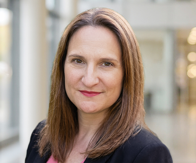 A woman with straight, shoulder-length brown hair and a light complexion is wearing a black blazer over a pink top. She is smiling slightly and standing in a bright, modern indoor setting with blurred background.