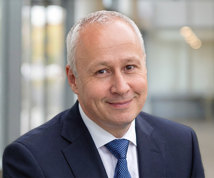 A middle-aged man with short grey hair wearing a dark suit, white shirt, and blue tie smiles gently at the camera in a bright, modern office setting.