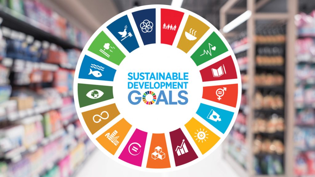 Circular graphic of the Sustainable Development Goals with icons for each goal, such as health, education, and climate action, over a blurred background of supermarket shelves.