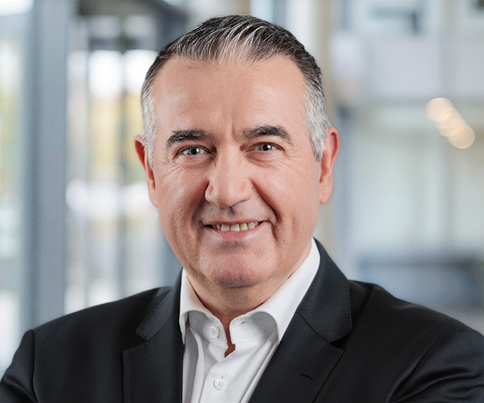 A middle-aged man with short grey hair smiles confidently whilst wearing a black suit jacket and white dress shirt, standing in a modern office environment with blurred background.