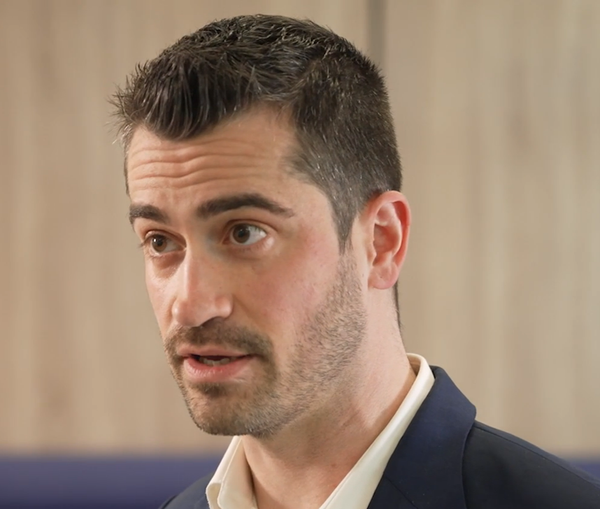 A man with short dark hair and a beard is seen talking. He is wearing a dark blazer over a light-colored shirt. The background is blurred, showing neutral-toned walls.