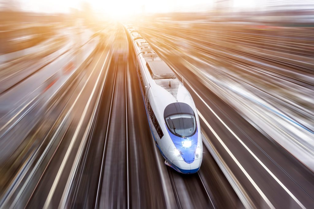 A sleek, modern high-speed train is moving rapidly along railway tracks, with a blurred background that emphasizes its speed. The sun is setting or rising in the distance, casting a warm golden light over the scene.