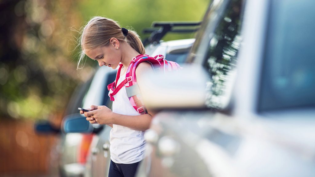 A young girl with a pink backpack stands between parked cars while looking down at her smartphone, appearing focused. The background is outdoors and slightly blurred, suggesting a parking lot or street.