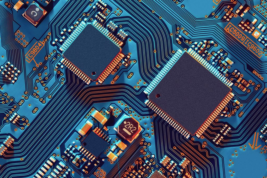 Close-up image of an electronic circuit board with two large microchips, numerous smaller components, and intricate blue and orange conductive pathways connecting them. The circuit board displays a complex and detailed pattern of interconnected technology.