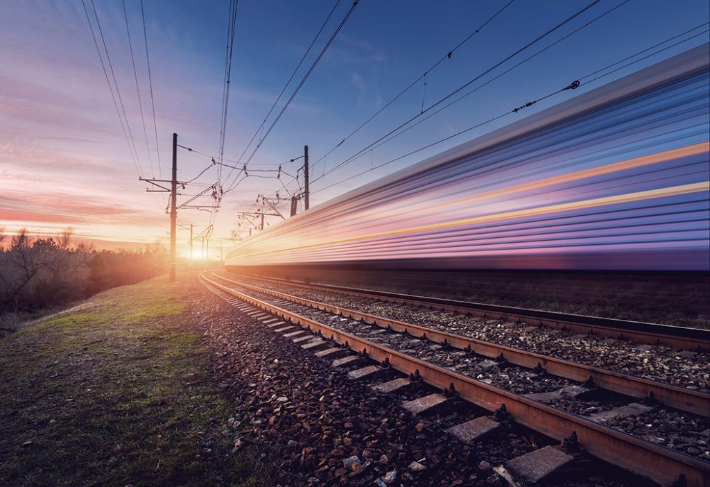 A train speeds along a track at sunset, creating a blurred effect. The sky transitions from a pink and orange sunset on the left to a clear blue on the right. Power lines run parallel to the tracks, and grassy terrain stretches to the left side of the image.
