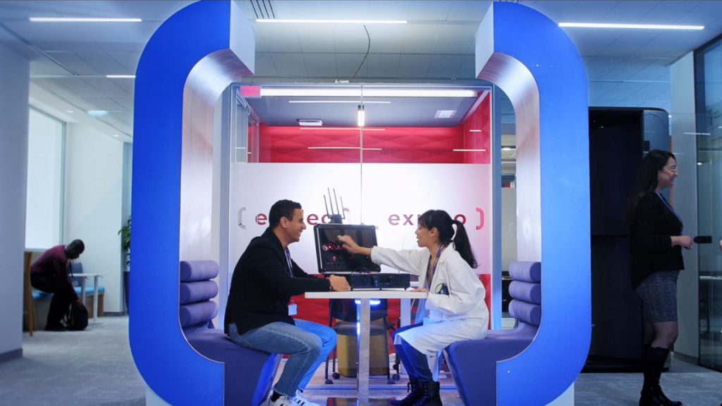 Two people are sitting and interacting at a small, enclosed workspace with blue arches. One is wearing a lab coat. In the background, a person sits typing on their laptop while another walks past. The workspace has soft lighting and modern design elements.