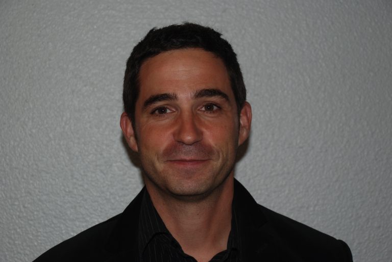 A man with short dark hair and a slight smile looks directly at the camera. He is wearing a dark suit jacket over a dark shirt. The background is a plain, textured light grey wall.