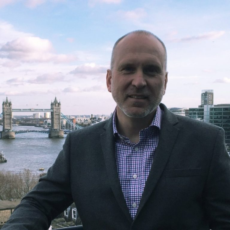 A man in a grey blazer and checkered shirt stands in front of a scenic view of Tower Bridge in London. The Thames River and modern city buildings, with hints of embedded software innovations, are visible in the background under a partly cloudy sky.