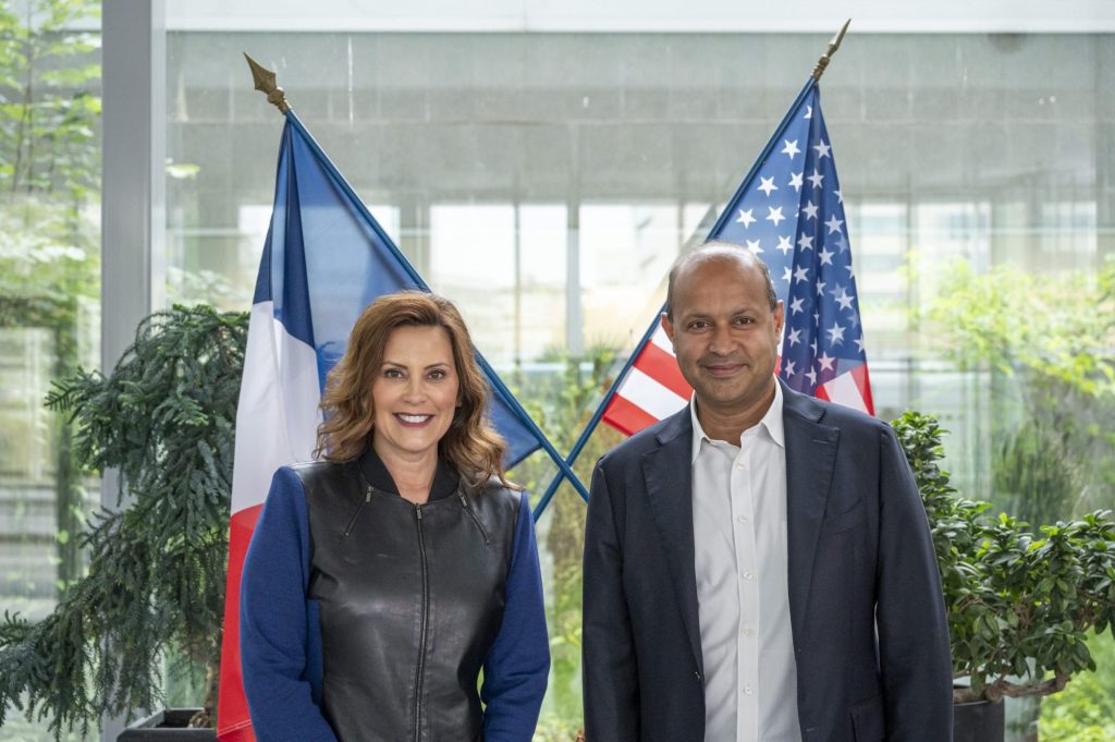 A woman and a man are standing side by side indoors with a glass window in the background. Both are smiling. Behind them, there are two flags on stands: the flag of France on the left and the flag of the United States on the right. Greenery is visible through the window.