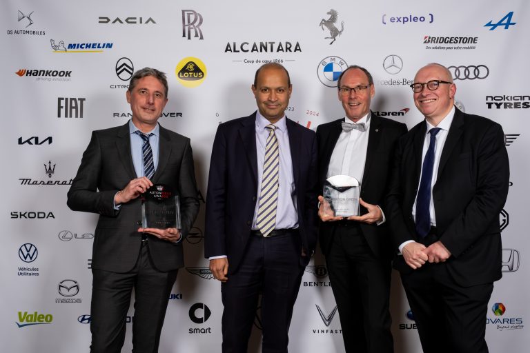 Four men in suits pose and smile on a red carpet, two holding glass trophies, in front of a backdrop featuring car manufacturer logos at the Innovationen Automobile Awards 2023.