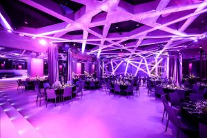 Modern event hall with geometric ceiling lights, purple ambient lighting, round tables set for a formal dinner, and elegantly decorated chairs. The space has a contemporary, stylish atmosphere.