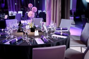 A round table set for a formal event with purple-themed decor, including napkins and floral centerpiece. Elegant glassware, plates, and silverware are neatly arranged under soft purple lighting.