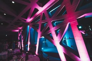 A modern interior with geometric wooden beams illuminated by pink and blue lights, creating an abstract, futuristic atmosphere. Tables with white tablecloths and glasses are set in the foreground.