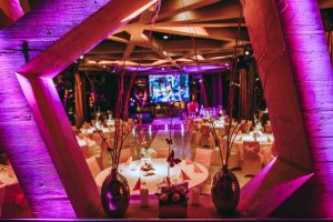 A modern event venue decorated with pink and purple lighting, featuring round tables with white tablecloths and floral centerpieces, viewed through a large geometric wooden frame.
