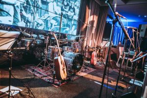A stage setup for a live performance featuring a drum set, two acoustic guitars, microphones, music stands, and various other musical equipment, with blue lighting and a large screen in the background.