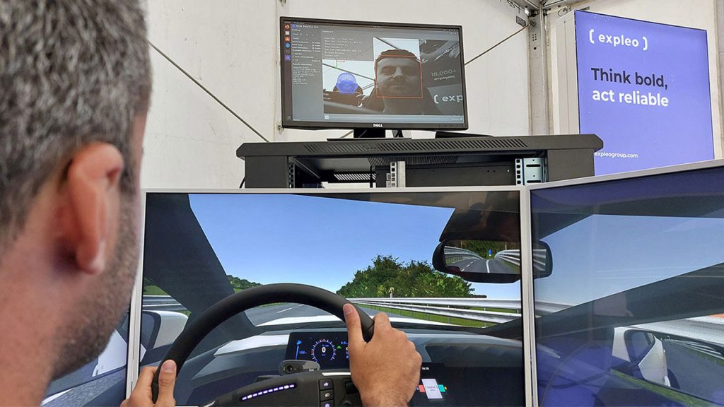 A person uses a driving simulator with three monitors showing a racetrack, while a screen above displays their face and simulation data. A sign nearby reads, "Think bold, act reliable.