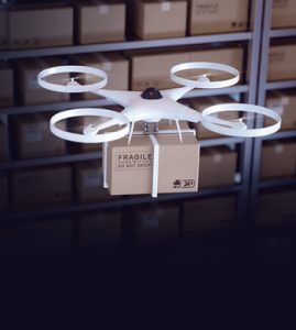 A drone with four propellers carries a cardboard box labeled "FRAGILE DO NOT DROP" in a warehouse, with shelves of stacked boxes in the background.