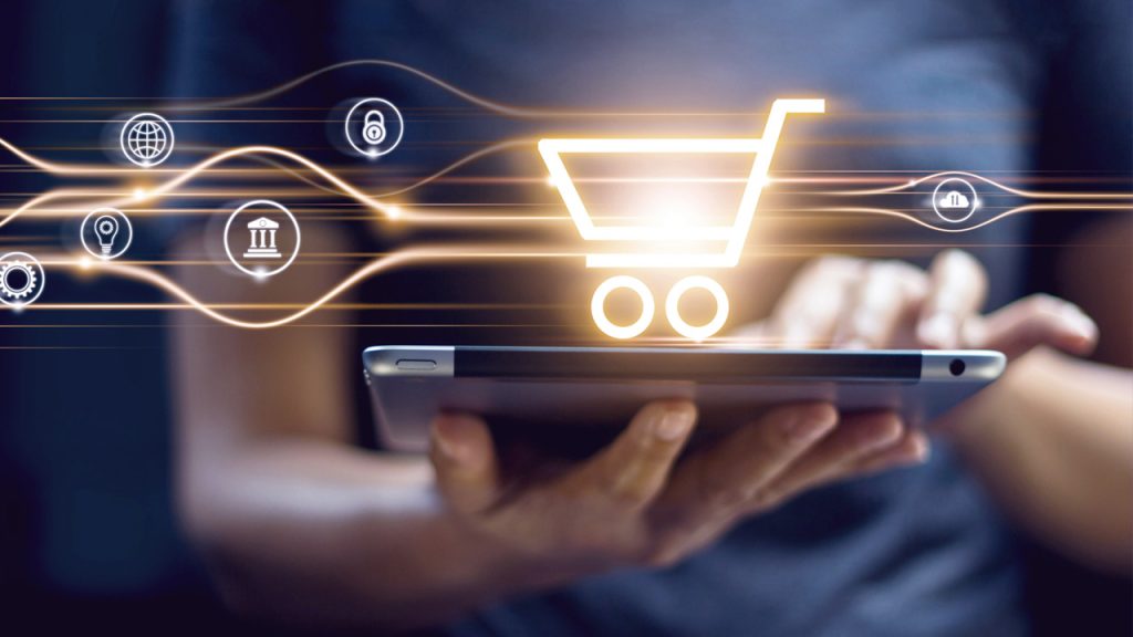 A person holds a tablet, with a glowing shopping cart icon hovering above the screen. Surrounding the icon are various symbols representing digital security and commerce, suggesting online shopping or e-commerce transactions.