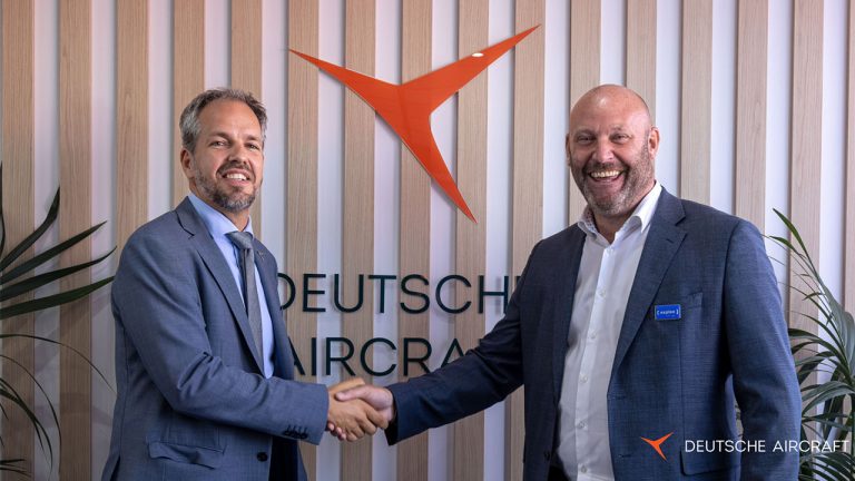 Two men in suits smile and shake hands in front of a wall with the Deutsche Aircraft logo and text. Vertical wooden slats and plants are visible in the background.