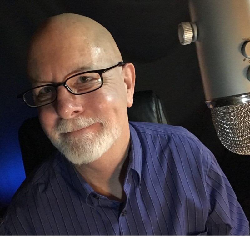 A bald man with glasses and a white beard, wearing a blue striped shirt, smiles at the camera whilst sitting near a large silver microphone. The background is dark.