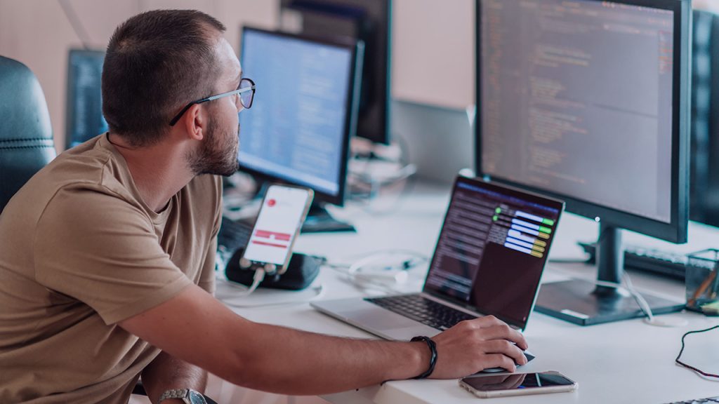A man wearing glasses sits at a desk, working on a computer with multiple monitors displaying code. He uses a mouse, and a smartphone and other electronics are on the desk.