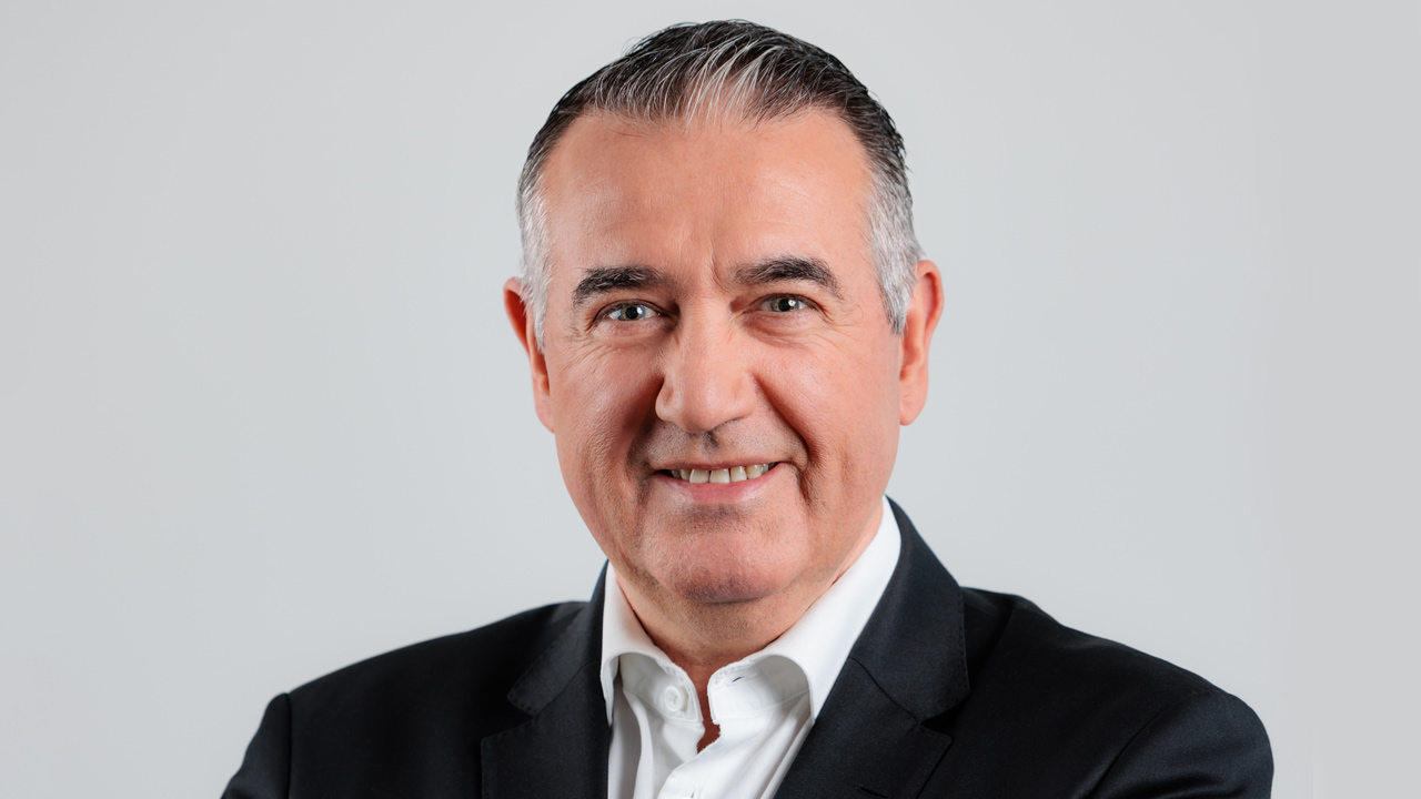 A middle-aged man with short, dark gray hair wearing a black suit jacket and white dress shirt, smiling warmly at the camera against a plain light gray background.