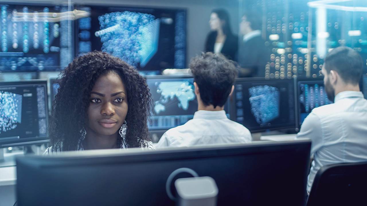 A woman sits at a computer in a modern office with multiple monitors displaying data and digital graphics, while two colleagues work at nearby desks and two people stand talking in the background.