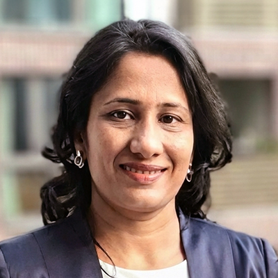 A woman with wavy dark hair smiles at the camera, wearing a navy blazer and silver earrings. The background is blurred, showing buildings and windows.