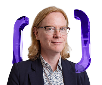 Person with shoulder-length blonde hair, glasses, patterned shirt, and dark blazer stands in front of a white background with large purple brackets on either side.