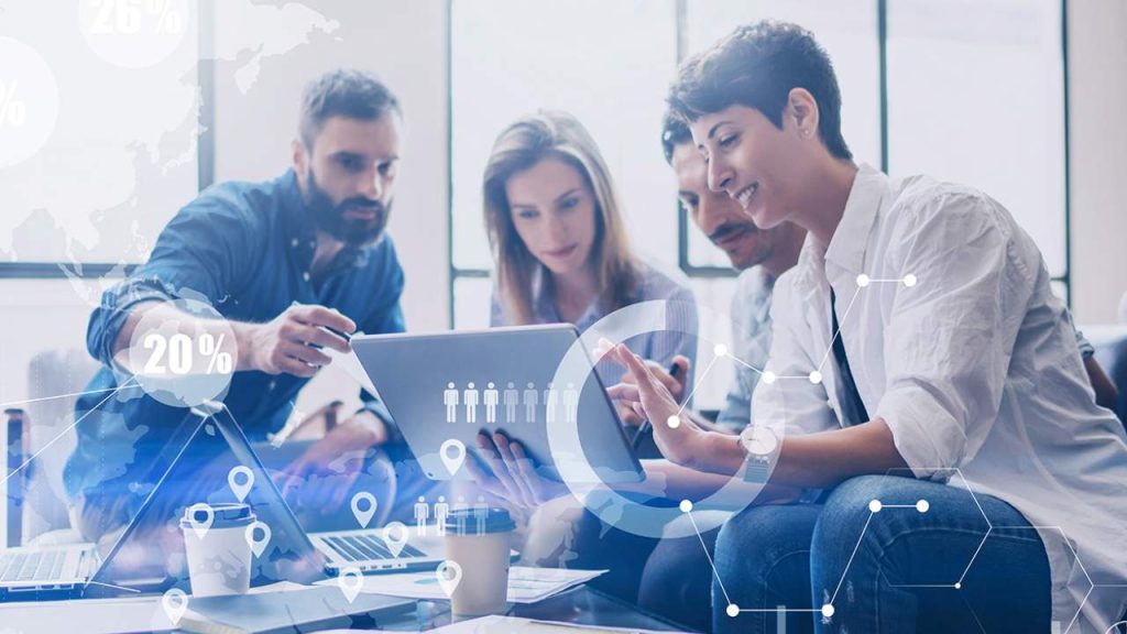 Four people sit at a table working together on laptops, discussing information displayed on a tablet. Transparent digital data graphics are layered over the image, illustrating technology and teamwork.