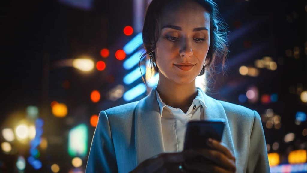 A woman in a light-coloured blazer looks at her mobile phone at night, with colourful city lights and blurred buildings in the background.