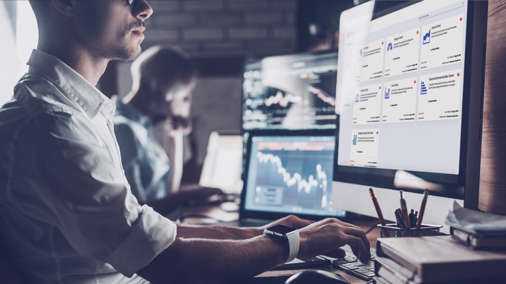 A person working at a desk with multiple computer monitors displaying graphs and data. A smartwatch is on their wrist. In the background, another person is also working at a desk with screens. The setting appears to be a modern office or trading room.