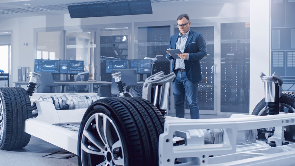 A man in business attire stands in a modern office, examining a digital tablet while looking at a car chassis prototype with exposed wheels and suspension, surrounded by computer monitors and glass walls.