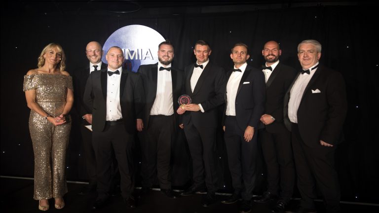A group of eight people, seven men in formal wear and one woman in a sequined dress, stand together on a stage. One man holds an award. They are against a backdrop with a circular logo.
