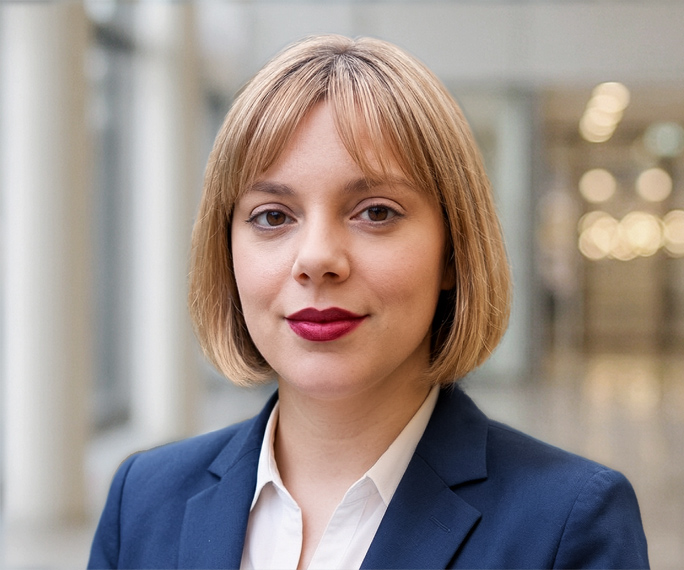 A woman with straight, blonde hair in a bob cut wears a navy blazer and white shirt, standing in a bright, modern indoor setting with blurred lights in the background.