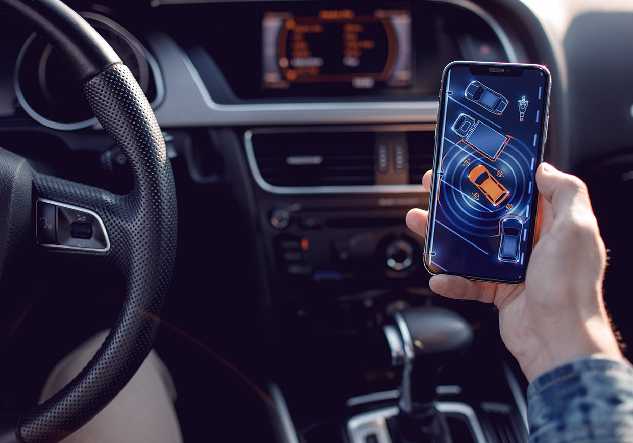 A person in a car holds a smartphone displaying a parking assist app, showing an overhead view of a car and nearby obstacles, while the car dashboard and steering wheel are visible.