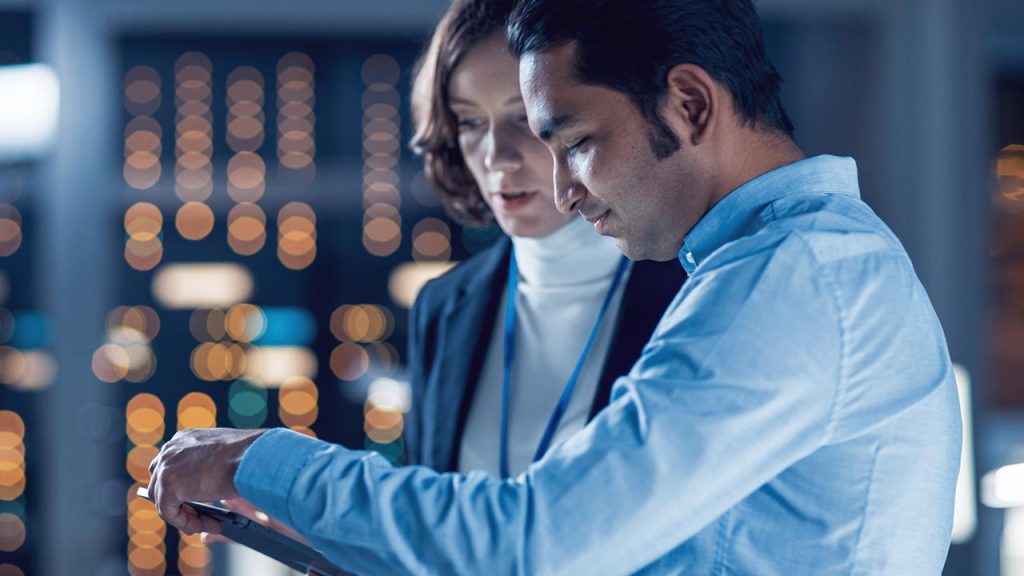 Two professionals, a man and a woman, closely looking at a tablet in a modern office setting with blurred lights and equipment in the background, suggesting a technology or data-focused environment.
