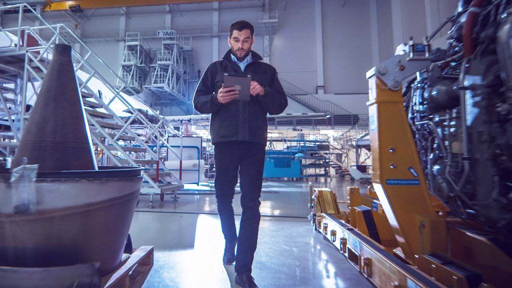 A man in a jacket walks through a brightly lit industrial facility, looking at a tablet. Manufacturing equipment and machinery are visible around him, and large metal structures fill the background.