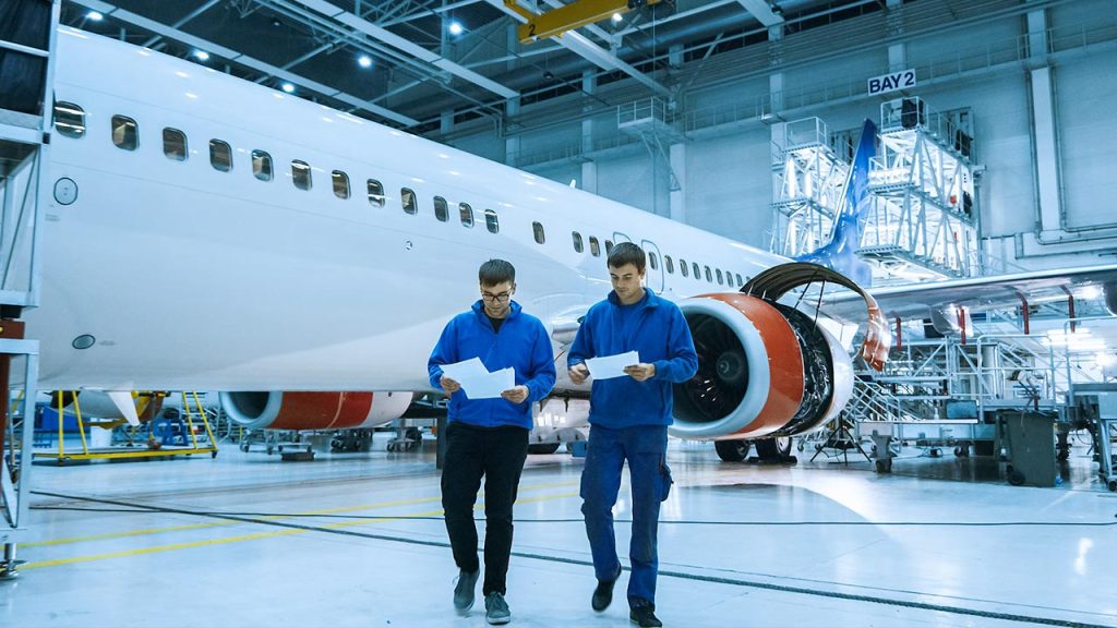 Two engineers in blue uniforms walk inside an aircraft hangar, holding papers and inspecting a large white airplane with red engine covers. The hangar is brightly lit and filled with aviation equipment.