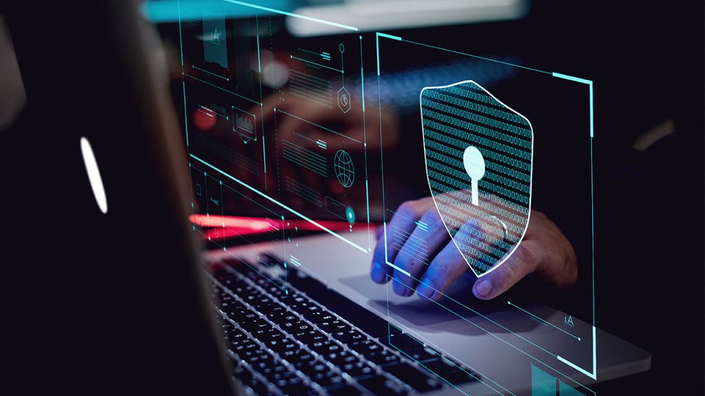 A person’s hand is using a laptop with digital security graphics overlaid, including a shield with a keyhole symbol, representing cybersecurity and data protection.