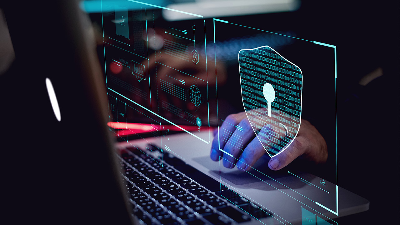 A person’s hand is using a laptop with digital security graphics overlaid, including a shield with a keyhole symbol, representing cybersecurity and data protection.