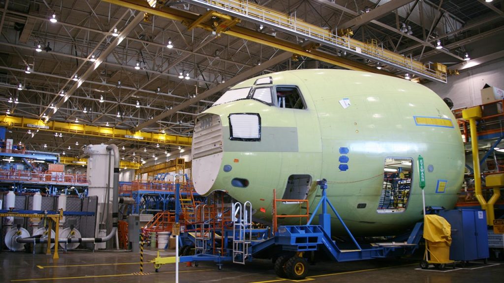 A large, unfinished airplane fuselage sits on a platform inside a spacious, well-lit aircraft manufacturing facility, surrounded by industrial equipment, scaffolding, and yellow overhead cranes.