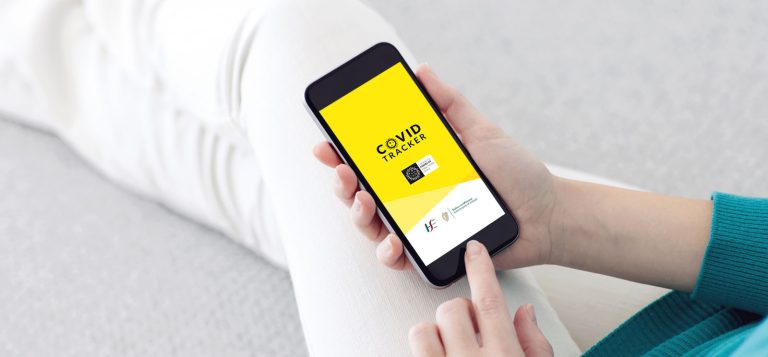 A person is holding a smartphone displaying a yellow screen with the "COVID Tracker" app open. The screen shows a QR code and logos of health organizations. The person is lying down on a couch and wearing a teal sweater.