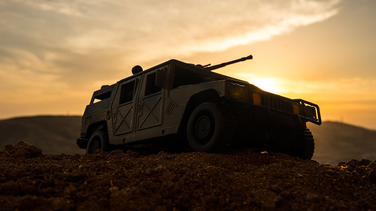 A military vehicle with a mounted gun is parked on rough terrain at sunset, silhouetted against a dramatic, cloudy sky with hills in the background.