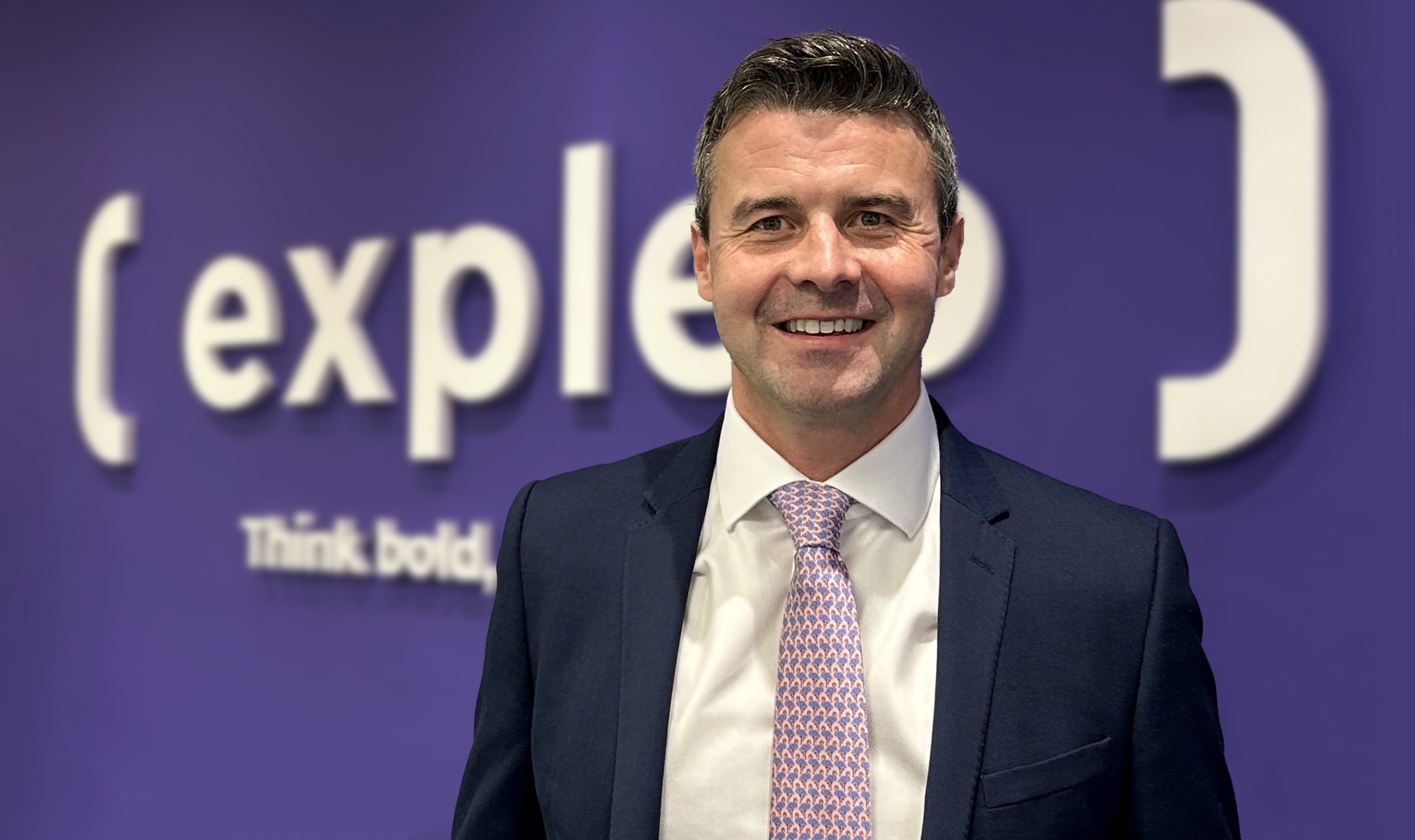 A man in a suit and patterned tie smiles at the camera, standing in front of a purple wall with the word “expleo” and the phrase “Think bold” partially visible in white text.